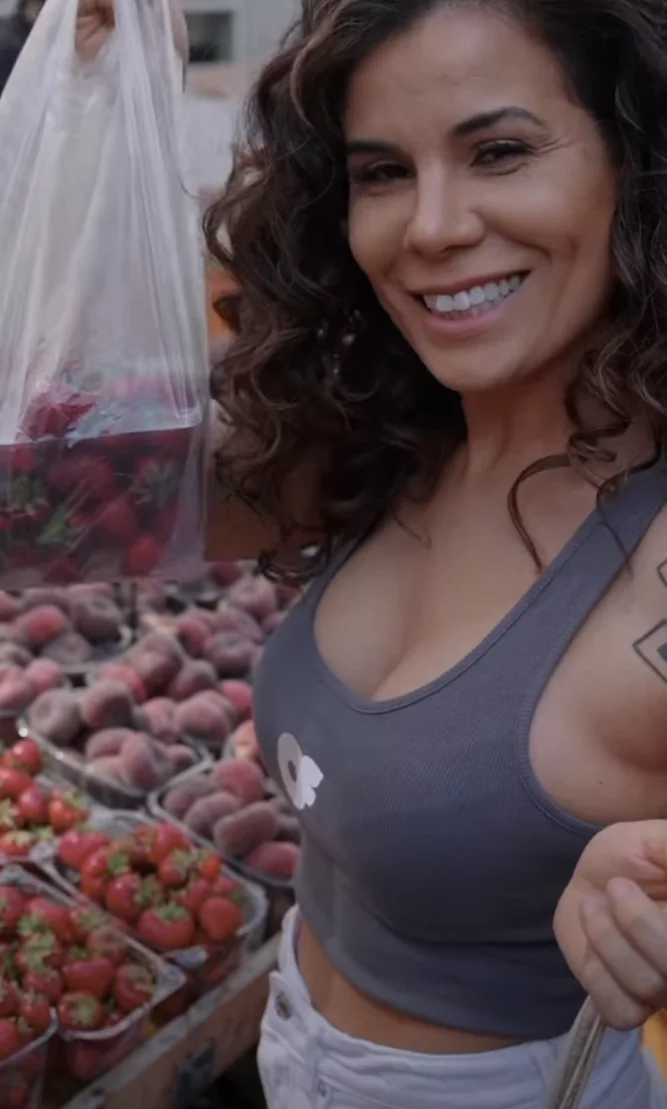 Cândida Batista num mercado, junto a bancas de fruta, com um saco de morangos na mão